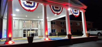 Entrada de un hotel con banderas estadounidenses, cortinas rojas y blancas, y un vehículo blanco estacionado afuera.