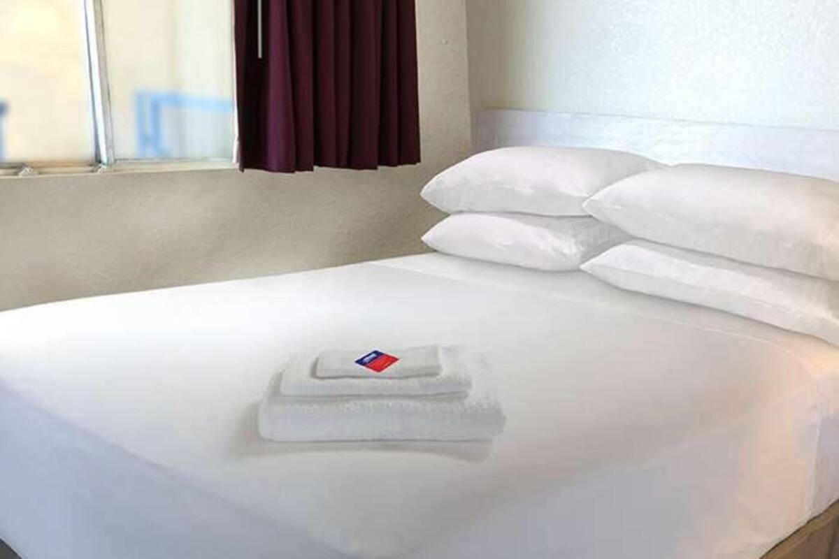A neatly made bed with white linens and a folded towel, a window with curtains, and a plain wall.