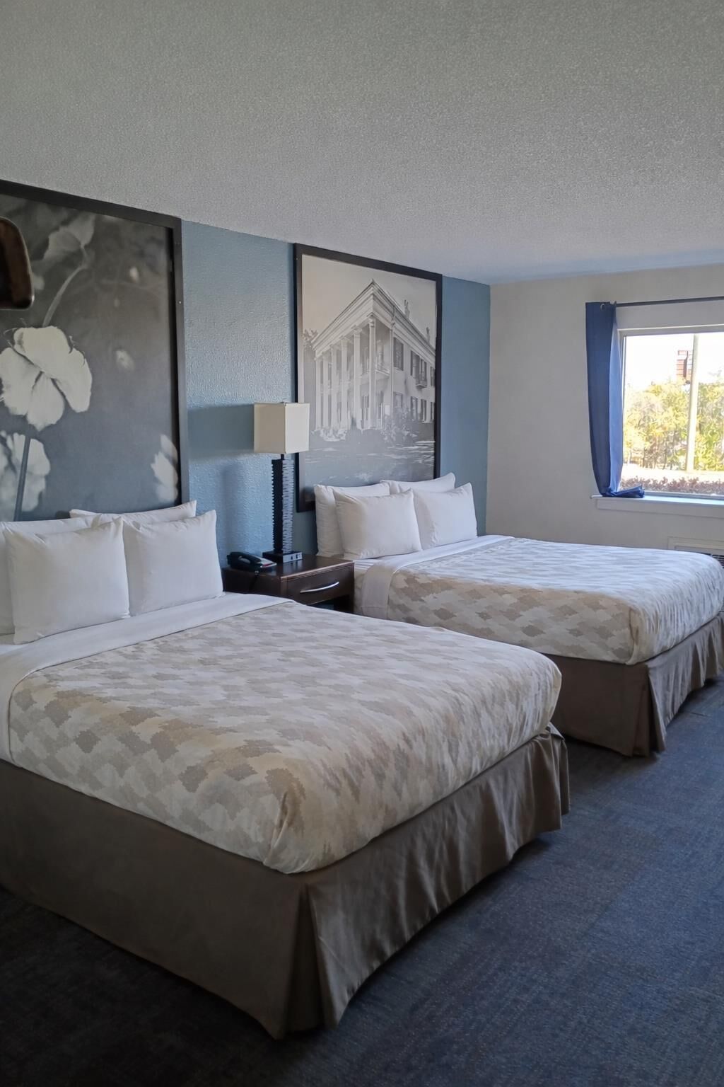 A hotel room with two beds, a nightstand, a lamp, and a large window with curtains.