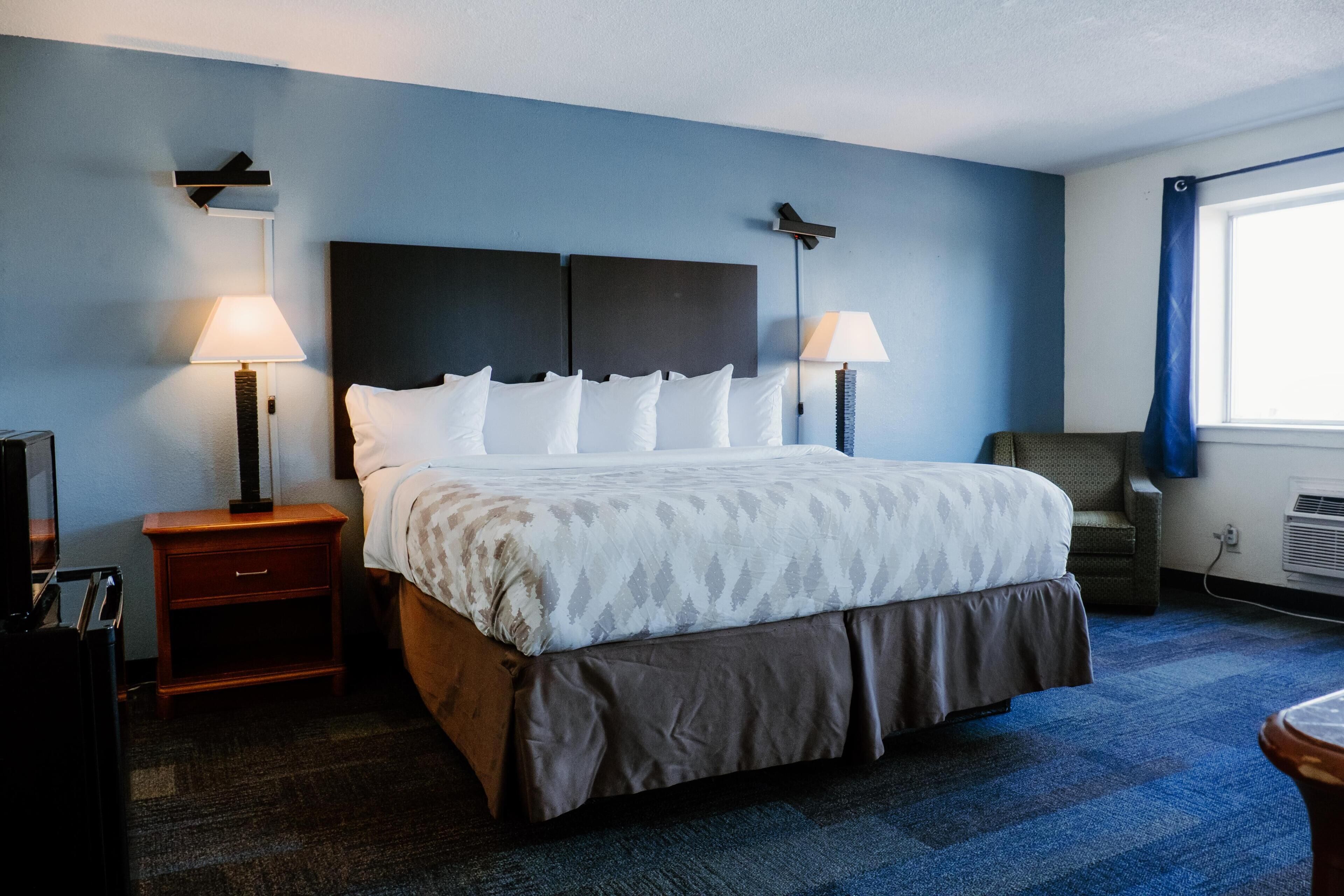 A hotel room with a large bed, two bedside lamps, a nightstand, a chair, and a window with curtains.