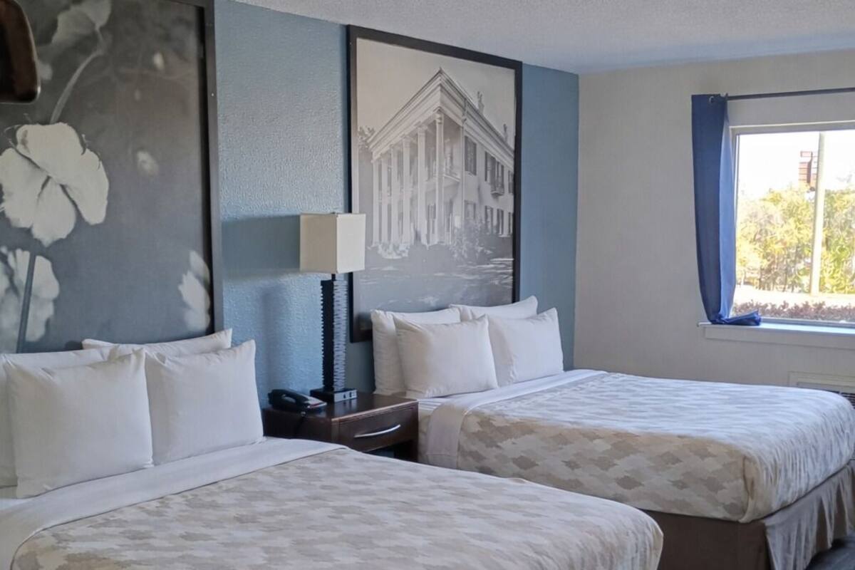 A hotel room with two beds, a large floral painting, a framed picture of a building, a nightstand, a lamp, and a window with curtains.