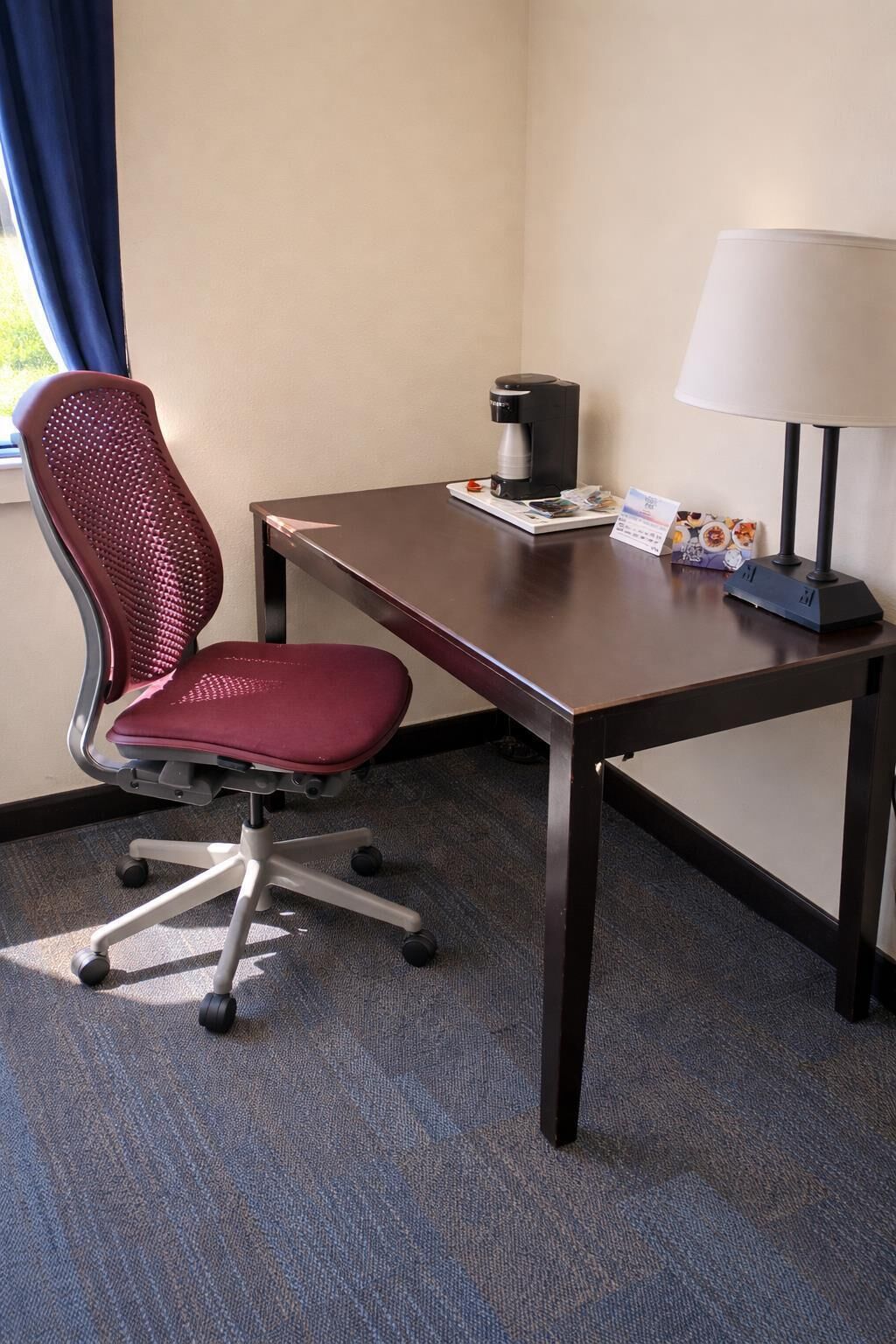 A hotel room with a desk, a chair, a coffee maker, a lamp, and a window with curtains.