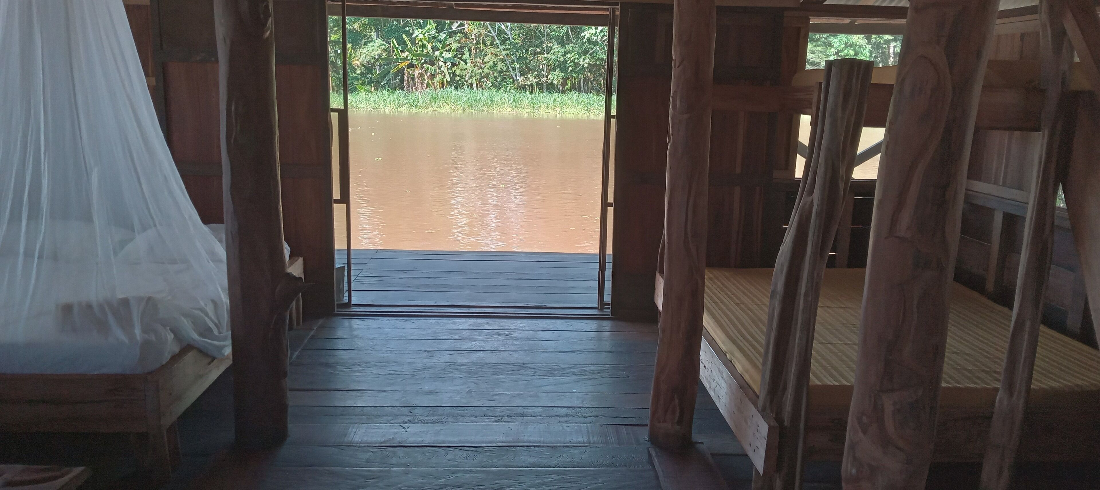 A wooden corridor leading to a water body, with a bed on the left and a wooden structure on the right.