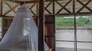 A wooden bed with a mosquito net, large windows, and a view of a river. - Kurupira Cabin Floating (Leticia)