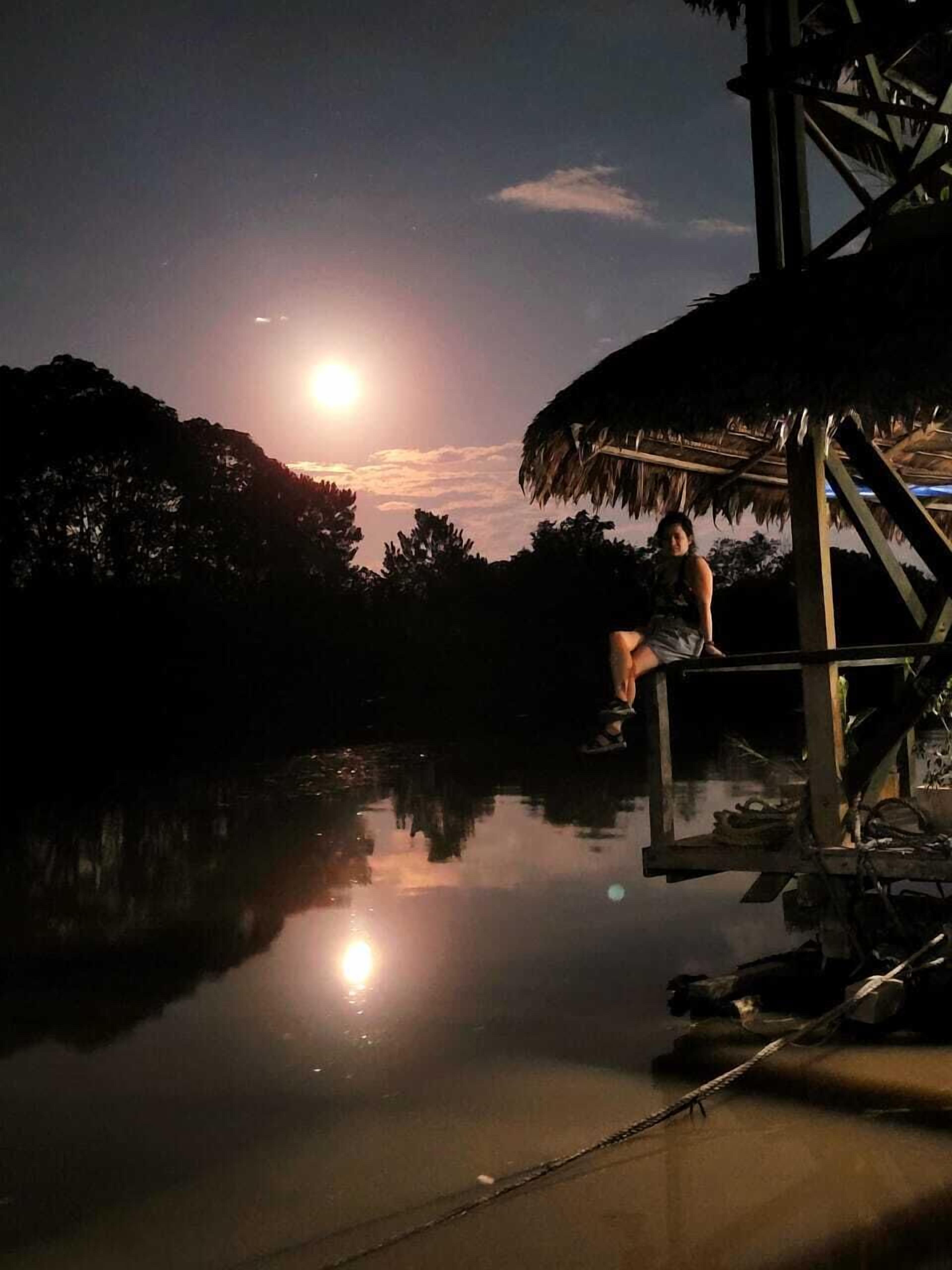 En person sidder på en trækonstruktion ved stille vand om natten, med måne og stjerners refleksioner i vandet.