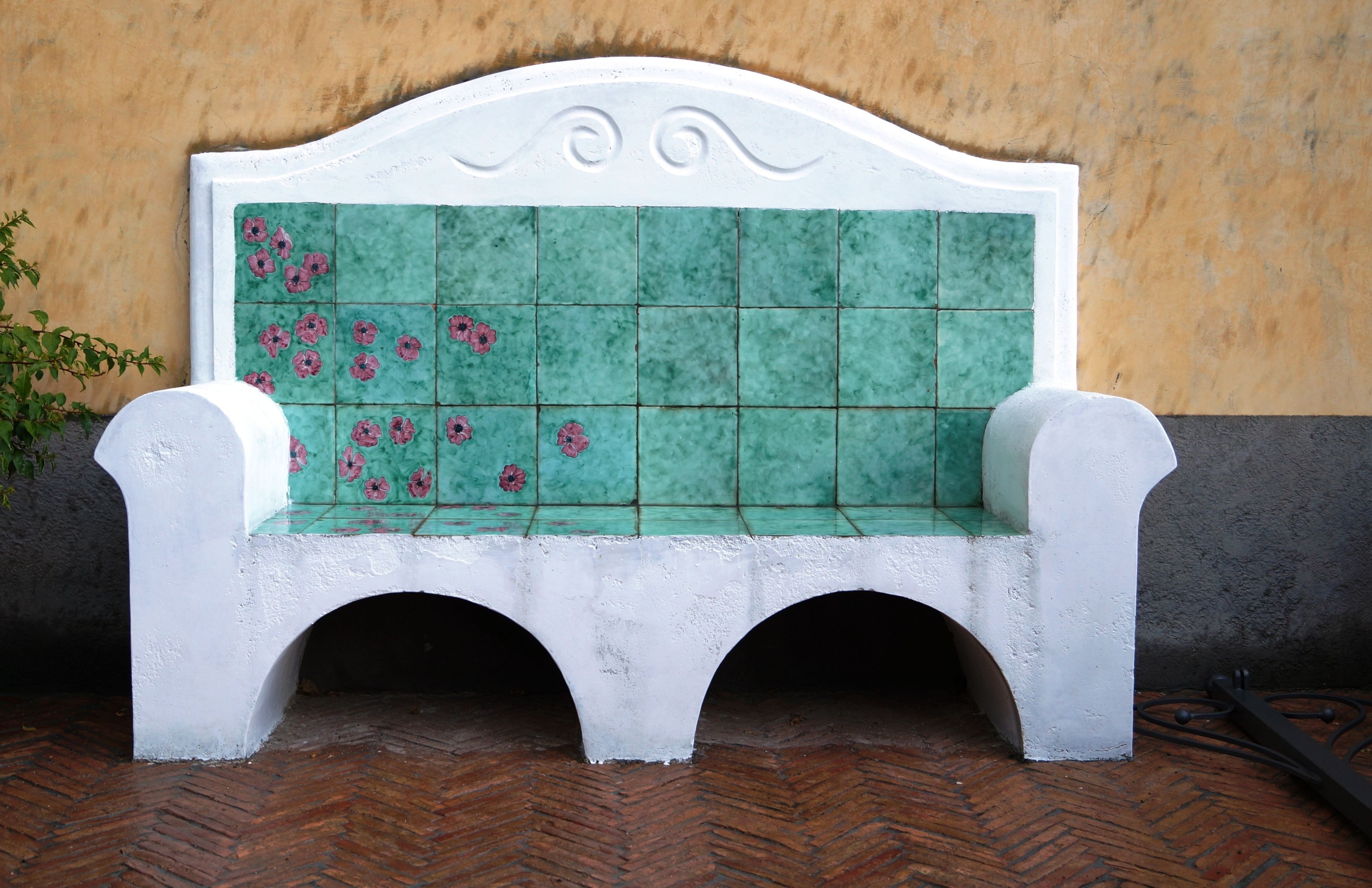 A white bench with a green tiled backrest and floral patterns, placed against a textured wall and wooden floor.