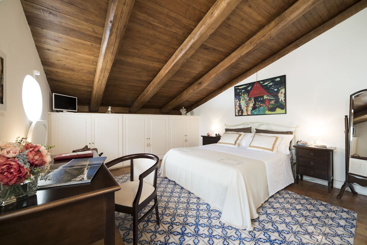 Ein Schlafzimmer mit Holzboden, einem Bett mit weißer Bettwäsche, einem Nachttisch, einem Sessel, einem Schreibtisch mit Blumen in einer Vase und einem Gemälde an der Wand.