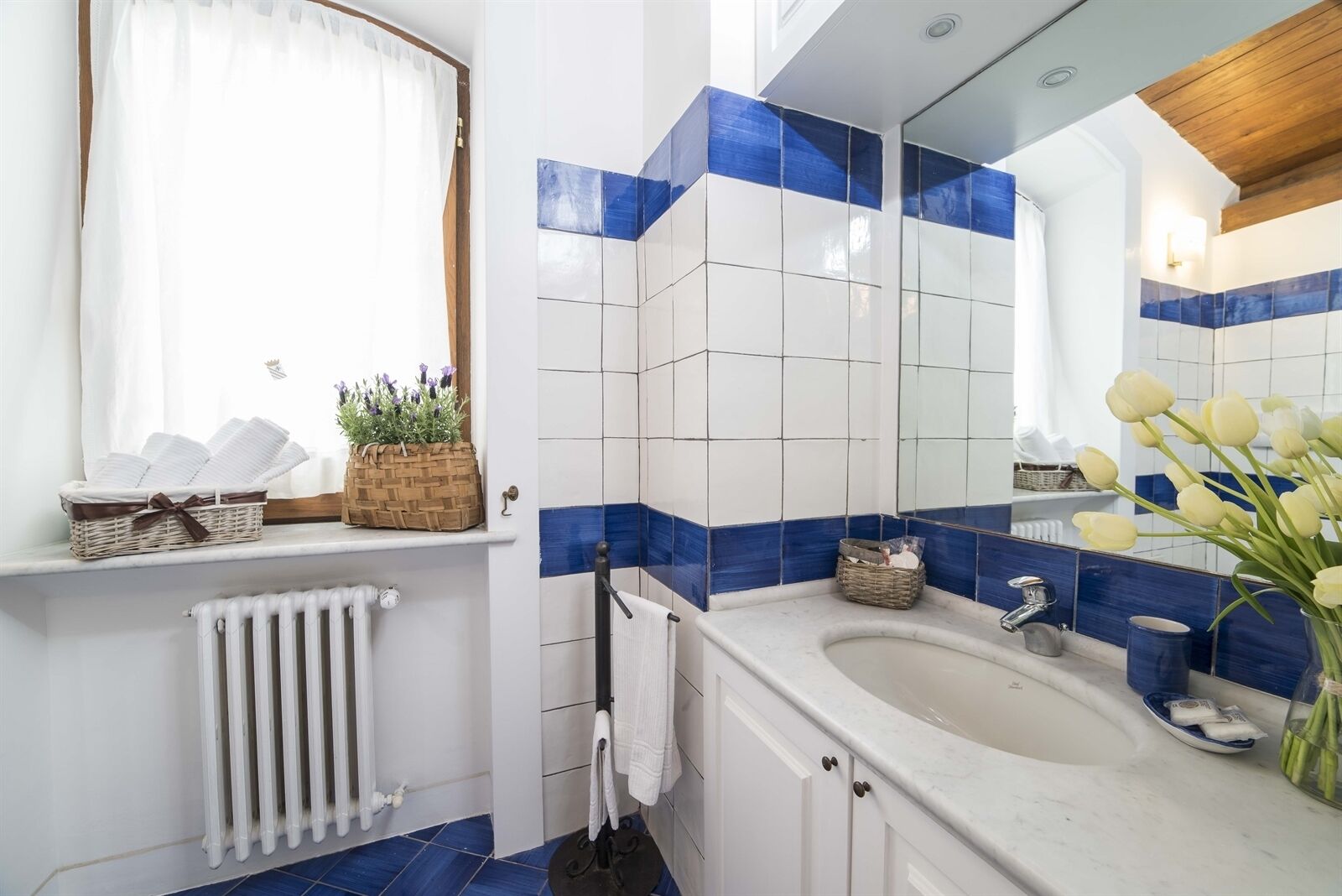 Un baño moderno con un lavamanos blanco, azulejos azules y blancos, una ventana con cortinas blancas y un espejo sobre el lavamanos.