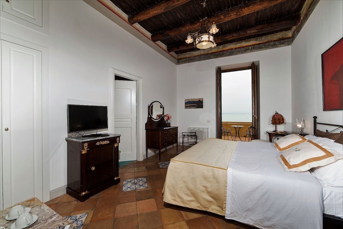 Ein Schlafzimmer mit Bett, Fernseher, Kommode, Stuhl und Meerblick durch ein Fenster.