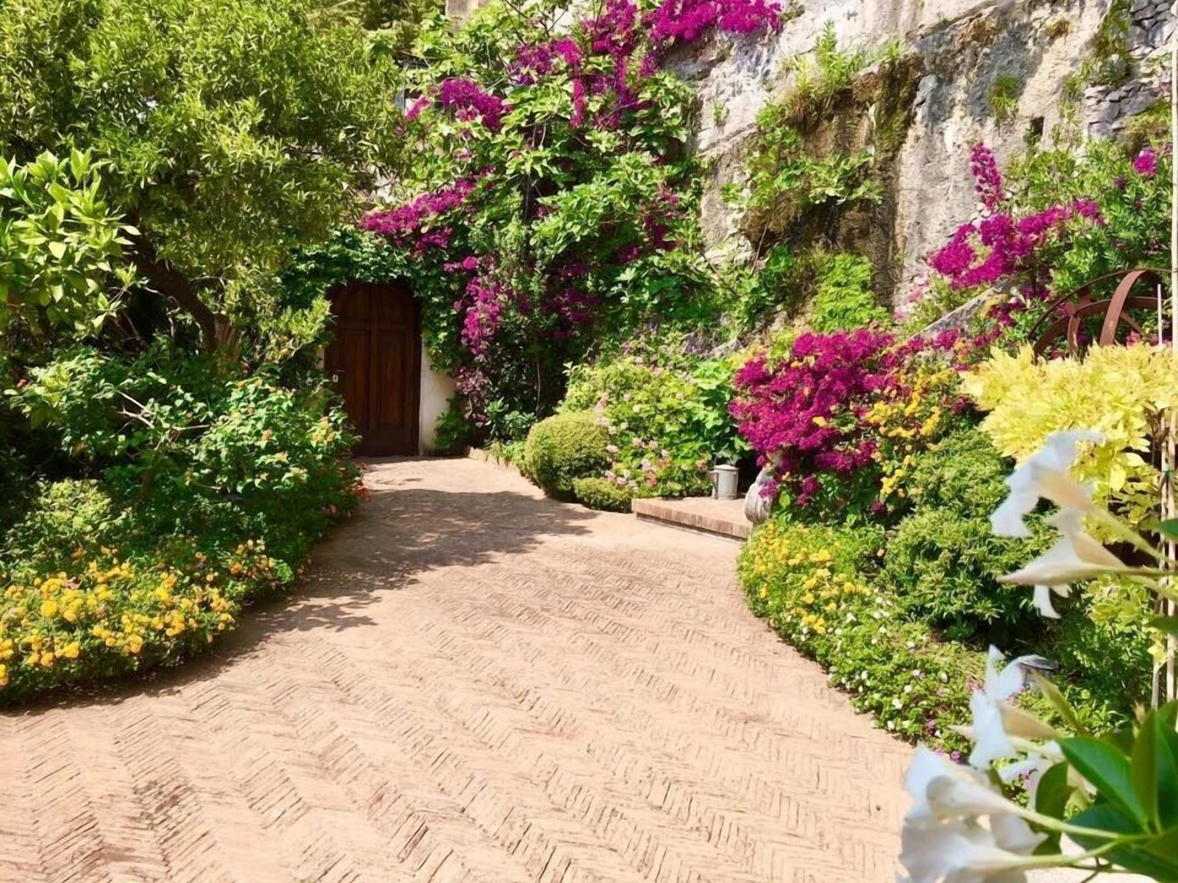 Un sendero pavimentado que conduce a una puerta de madera, rodeado de exuberante vegetación y flores coloridas.