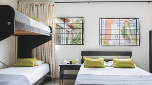 A hotel room with two beds, a bunk bed, a nightstand with a phone and a bottle, and two windows with views of buildings and trees. - Hotel Colonial Plaza (Bucaramanga)