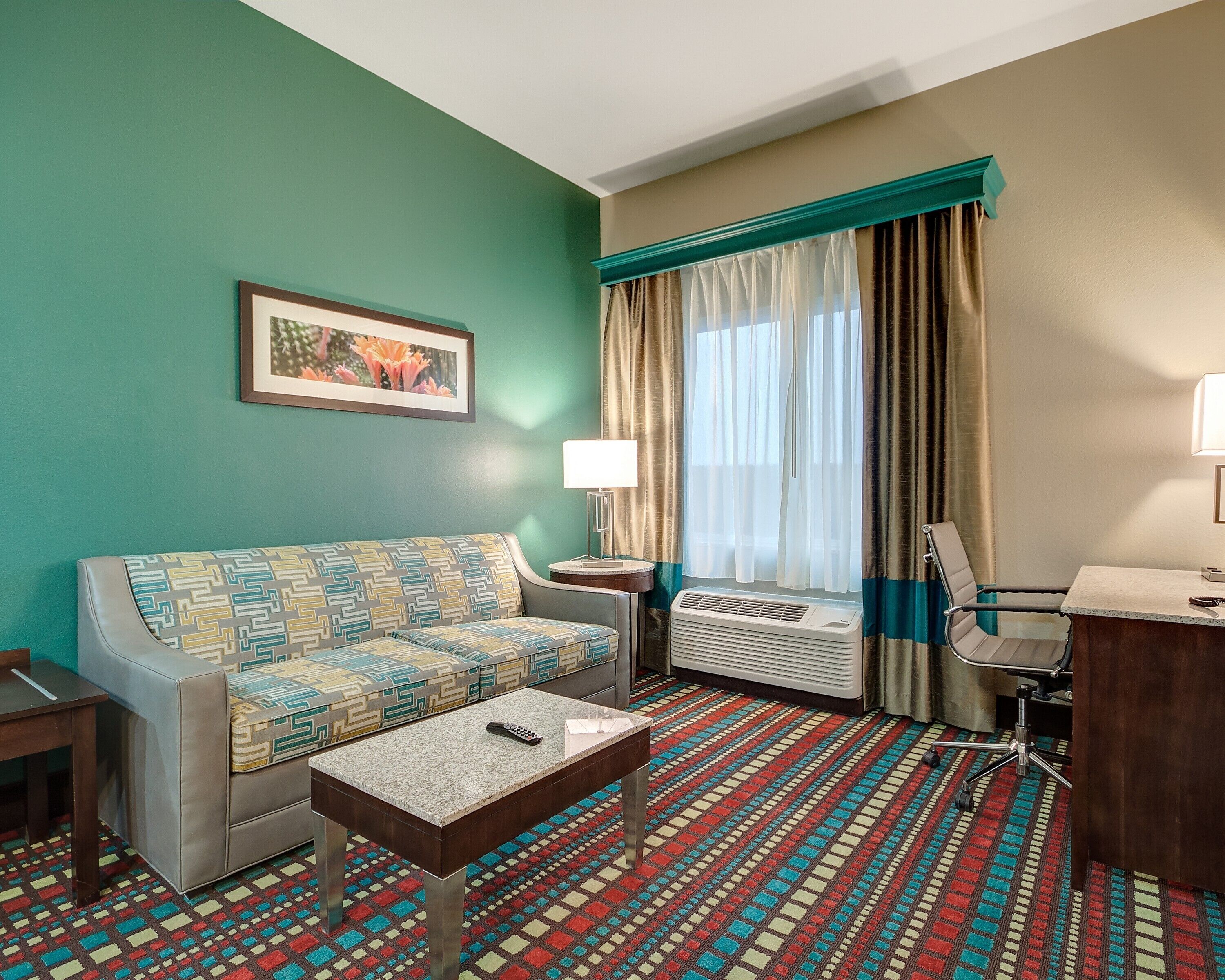 Habitación de hotel con sofá, mesa de centro, escritorio, silla, lámpara, aire acondicionado y ventana con cortinas.