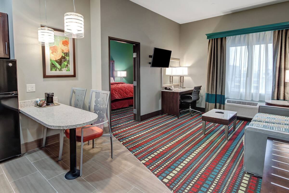 A hotel room with a kitchenette, a dining area, a bed with a red blanket, a desk with a chair, a TV, and a large window with curtains.