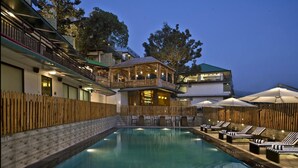 A pool area with lounge chairs and umbrellas, surrounded by wooden fences and buildings with balconies. - Fortune Park Moksha, Mcleodganj  - Member ITC Hotels' Group (Dharamshala)