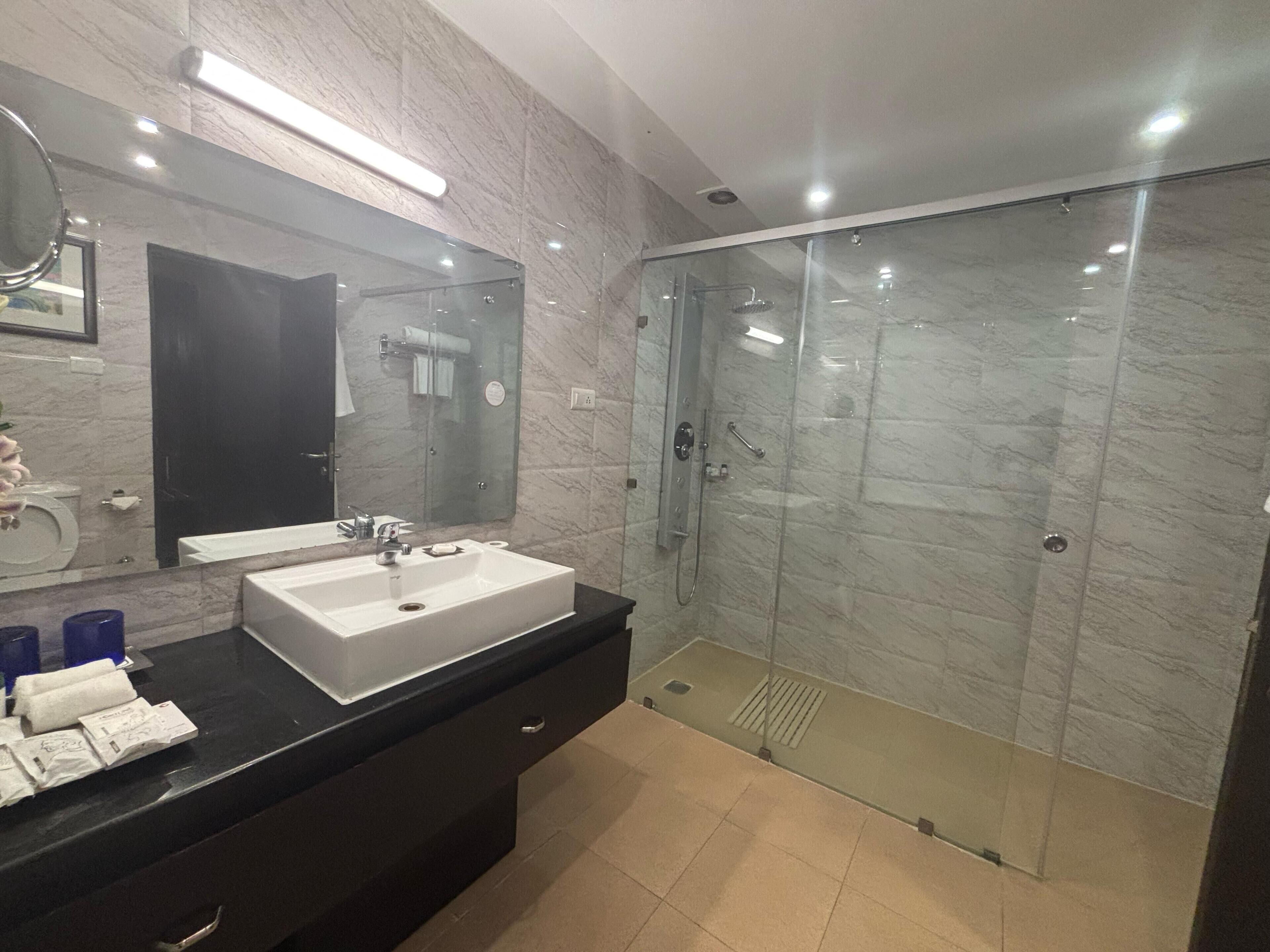 Un baño moderno con un espejo grande, un lavamanos blanco, una ducha con mampara de vidrio y un mueble de tocador negro.