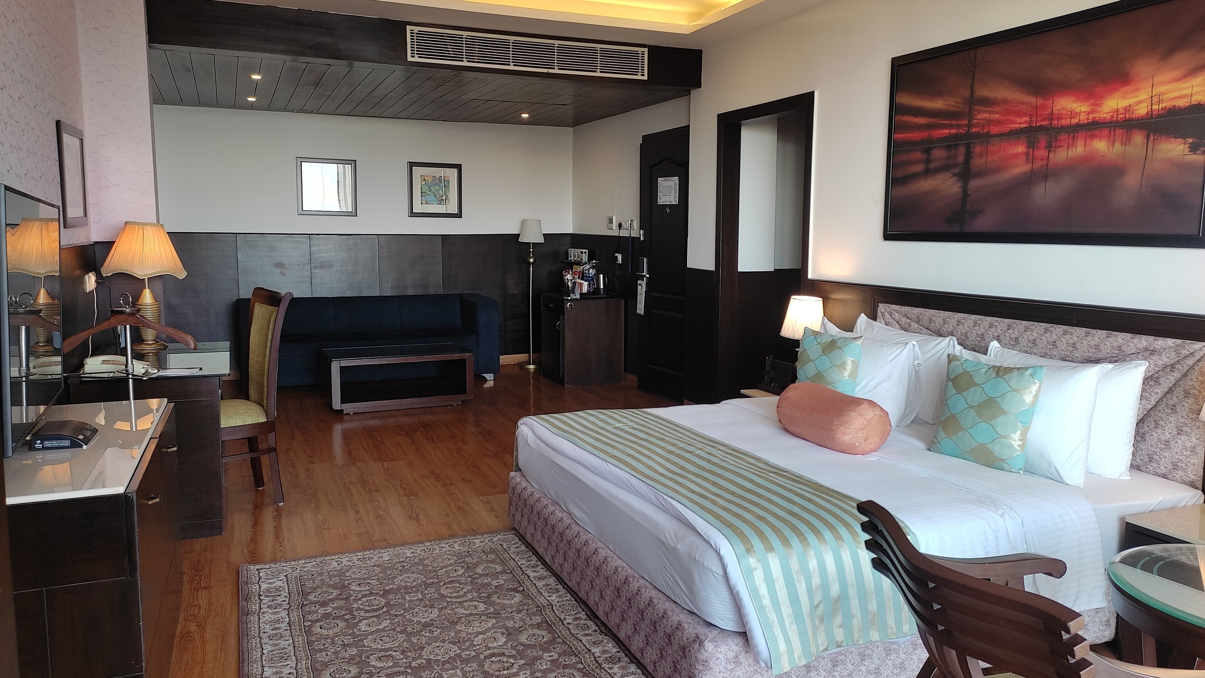 Una camera d'hotel con un letto, una scrivania, una sedia, un divano, una televisione e un quadro appeso al muro.