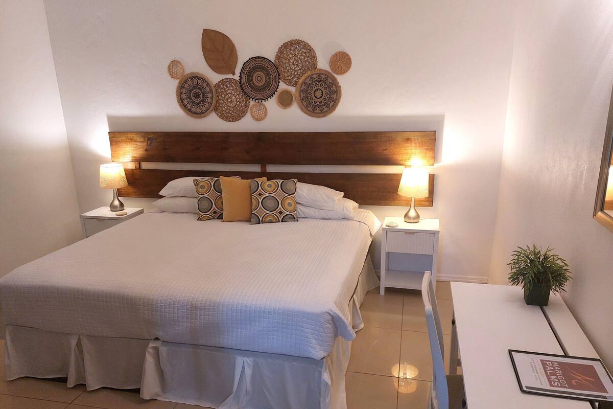 Un dormitorio ordenado con una cama grande, dos mesitas de noche con lámparas, un escritorio pequeño con una planta y decoración en la pared.