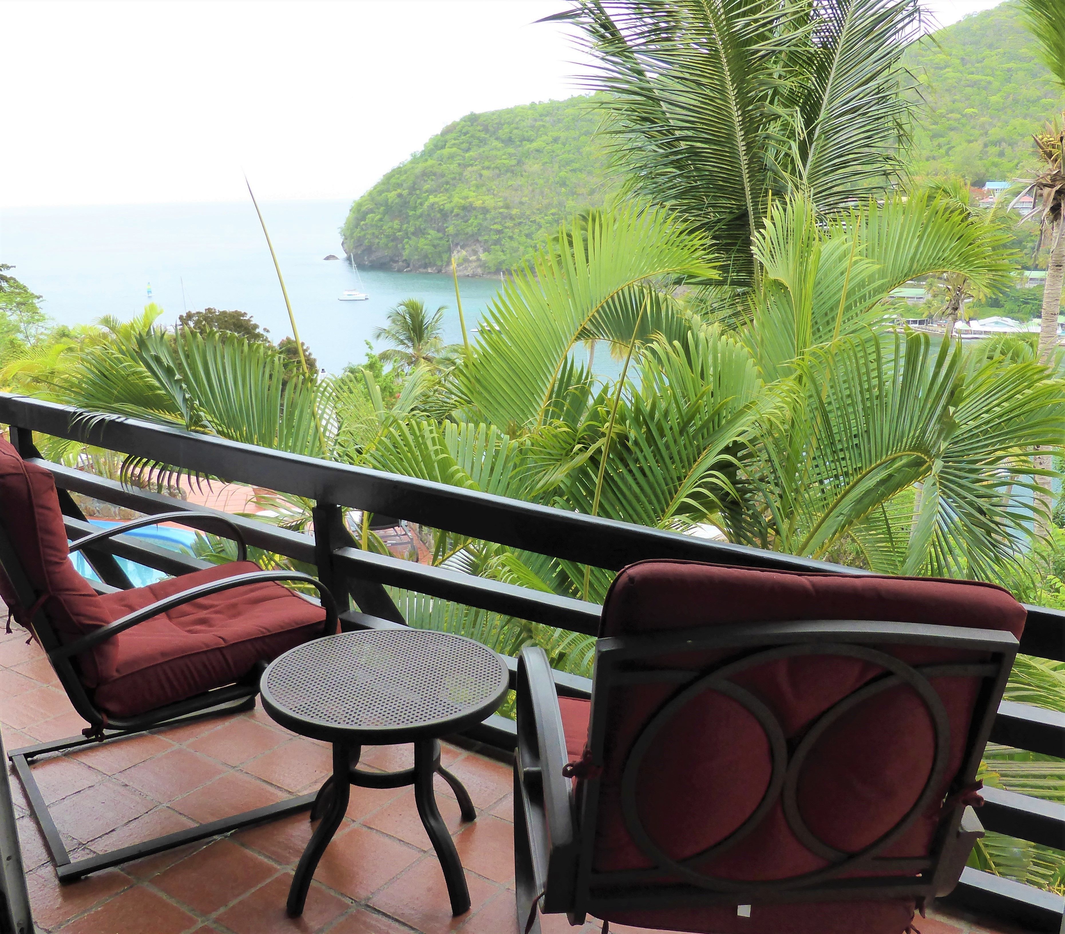 A balcony with two red cushioned chairs and a small round table, overlooking a coastal area with lush greenery and a view of the sea.
