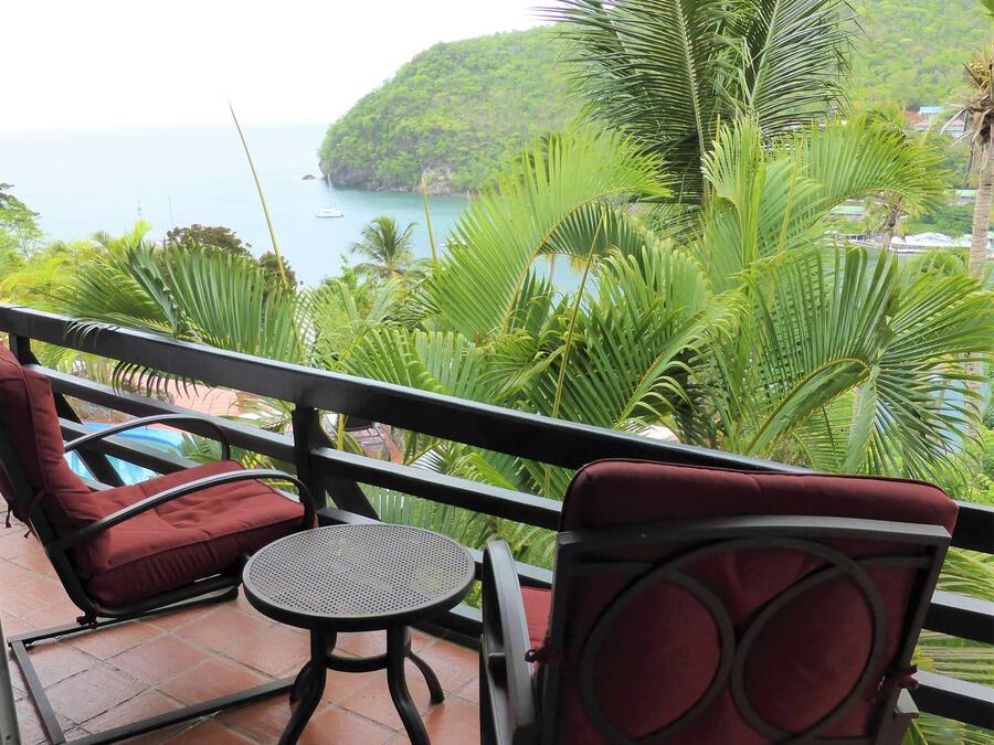 A balcony with two red cushioned chairs and a small round table, overlooking a coastal area with lush greenery and a view of the sea.