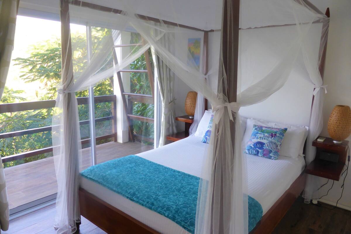 Un dormitorio con cama, mesita de noche, ventana con vista a árboles y balcón.