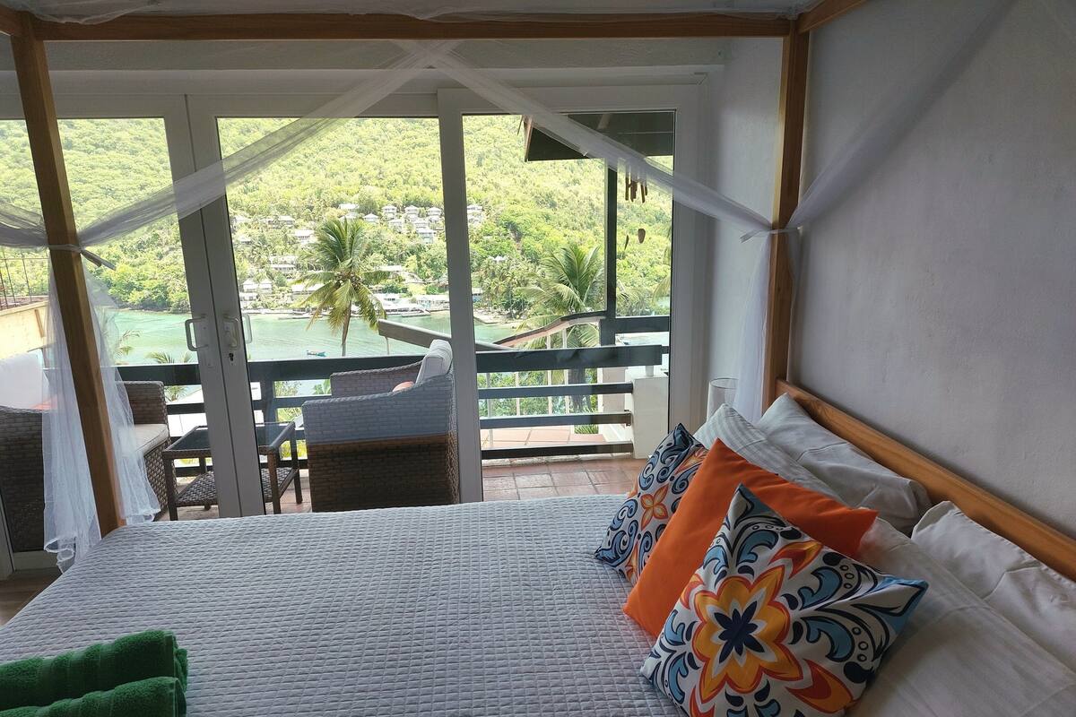 Un dormitorio con una cama grande, almohadas coloridas, una toalla verde y vistas a una piscina y follaje exterior.