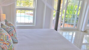 Un dormitorio con una cama grande, almohadones coloridos, un ventanal con vistas a una piscina y cortinas translúcidas.