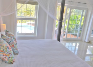 Une chambre à coucher avec un grand lit, des coussins aux couleurs vives, une fenêtre donnant sur une piscine et des rideaux légers.
