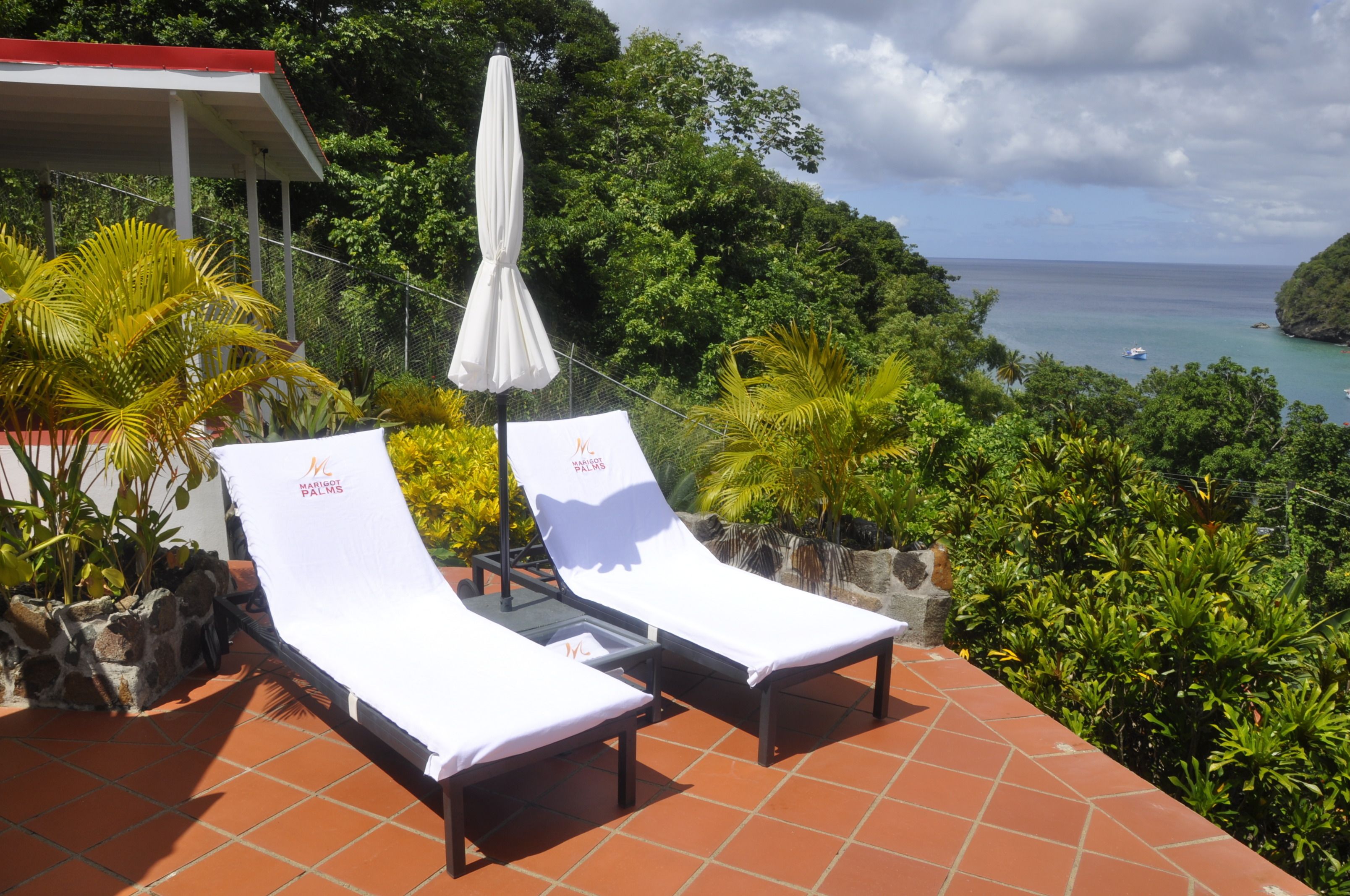 Two sun loungers with white cushions and a white umbrella, set on a tiled patio with a view of a tropical landscape and the sea.
