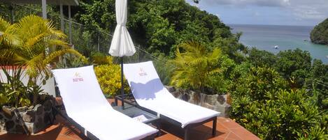Two sun loungers with white cushions and a white umbrella, set on a tiled patio with a view of a tropical landscape and the sea.