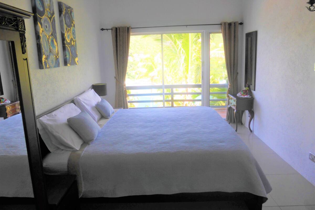 Un dormitorio con una cama grande, una mesita de noche, una lámpara y vistas a palmeras a través de la ventana.