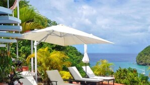 Zona junto a la piscina con sillones de descanso y una sombrilla grande, con vistas al mar azul turquesa y vegetación exuberante.
