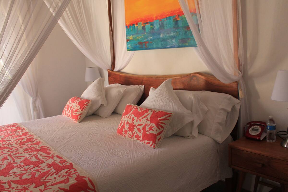 A neatly made bed with white and red floral pillows, a wooden headboard, and a bedside table with a lamp and a bottle.