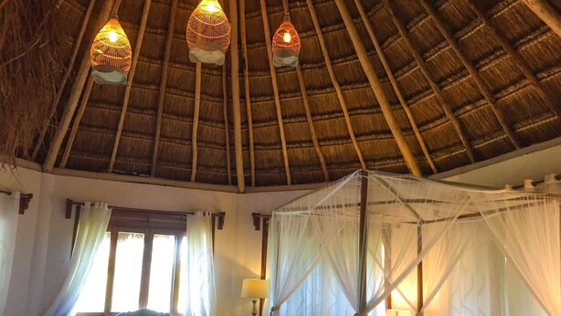 Una habitación con techo de paja, una cama con dosel, un sofá con cojines y una ventana con cortinas.