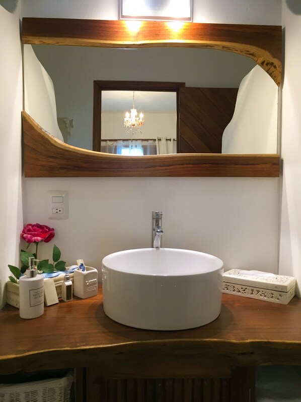 Un baño con un lavamanos blanco y redondo, un mueble de tocador de madera, un espejo grande y una araña.