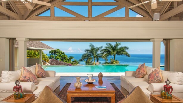 Une chambre luxueuse dotée d’une grande fenêtre donnant sur une piscine et l’océan. On y trouve un canapé blanc agrémenté de coussins colorés et une table basse en bois placée au centre de la pièce.