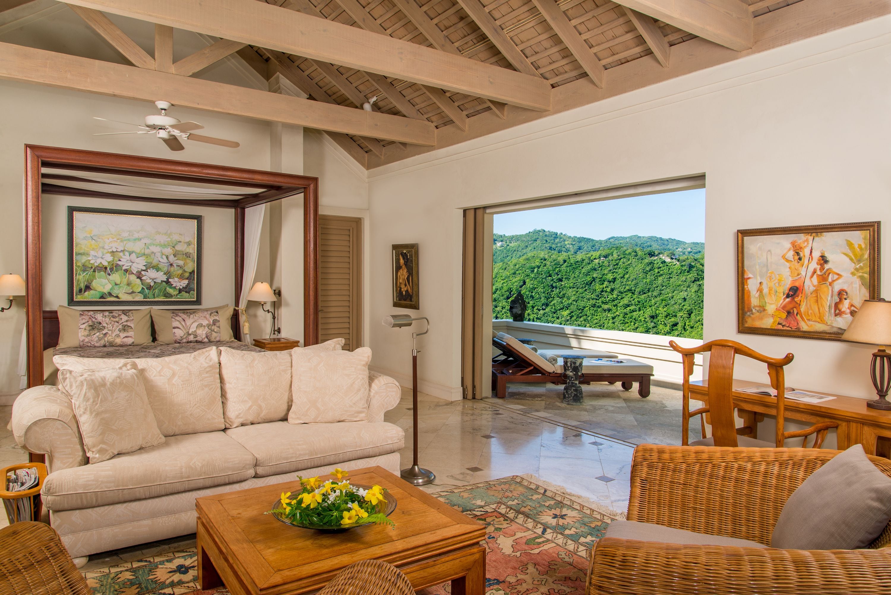 A spacious living room with a large window offering a view of lush greenery. A comfortable sofa and a wooden coffee table with flowers are in the foreground.