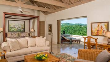 A spacious living room with a large window offering a view of lush greenery. A comfortable sofa and a wooden coffee table with flowers are in the foreground.
