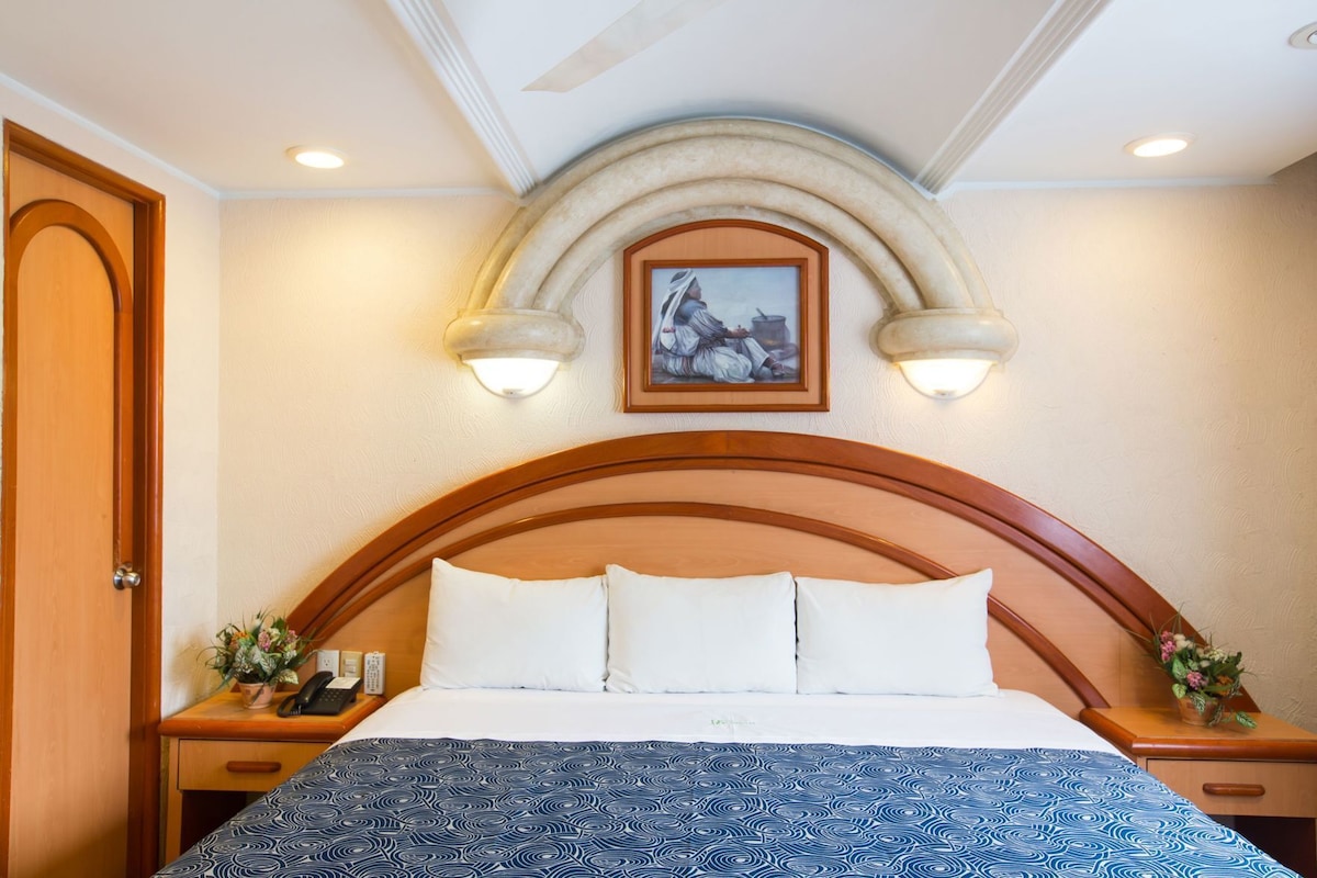 A hotel room with a large bed, a wooden headboard, two bedside tables with lamps, a painting above the headboard, and a door on the left.