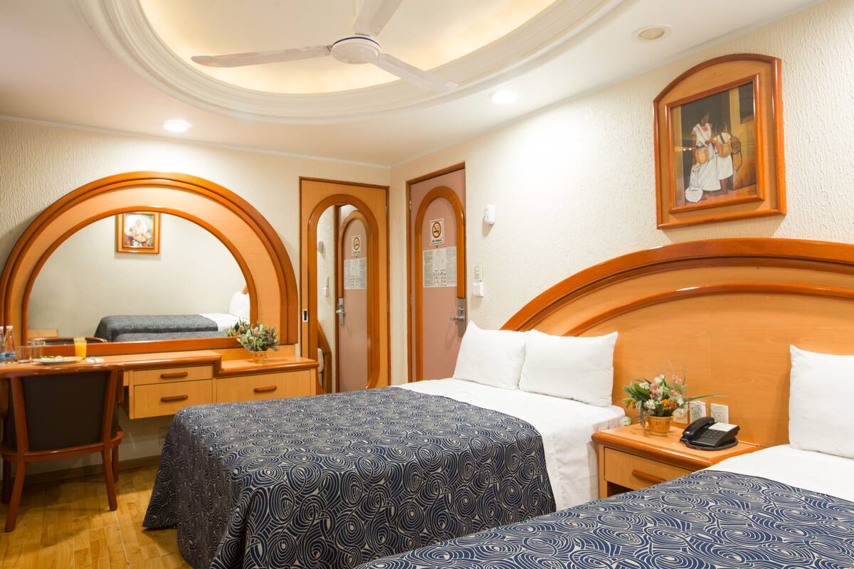 A hotel room with two beds, a desk, a chair, a mirror, and a ceiling fan.