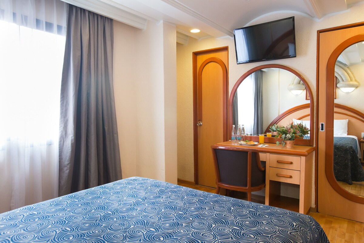 Una habitación de hotel con cama, escritorio, silla, espejo y televisión.