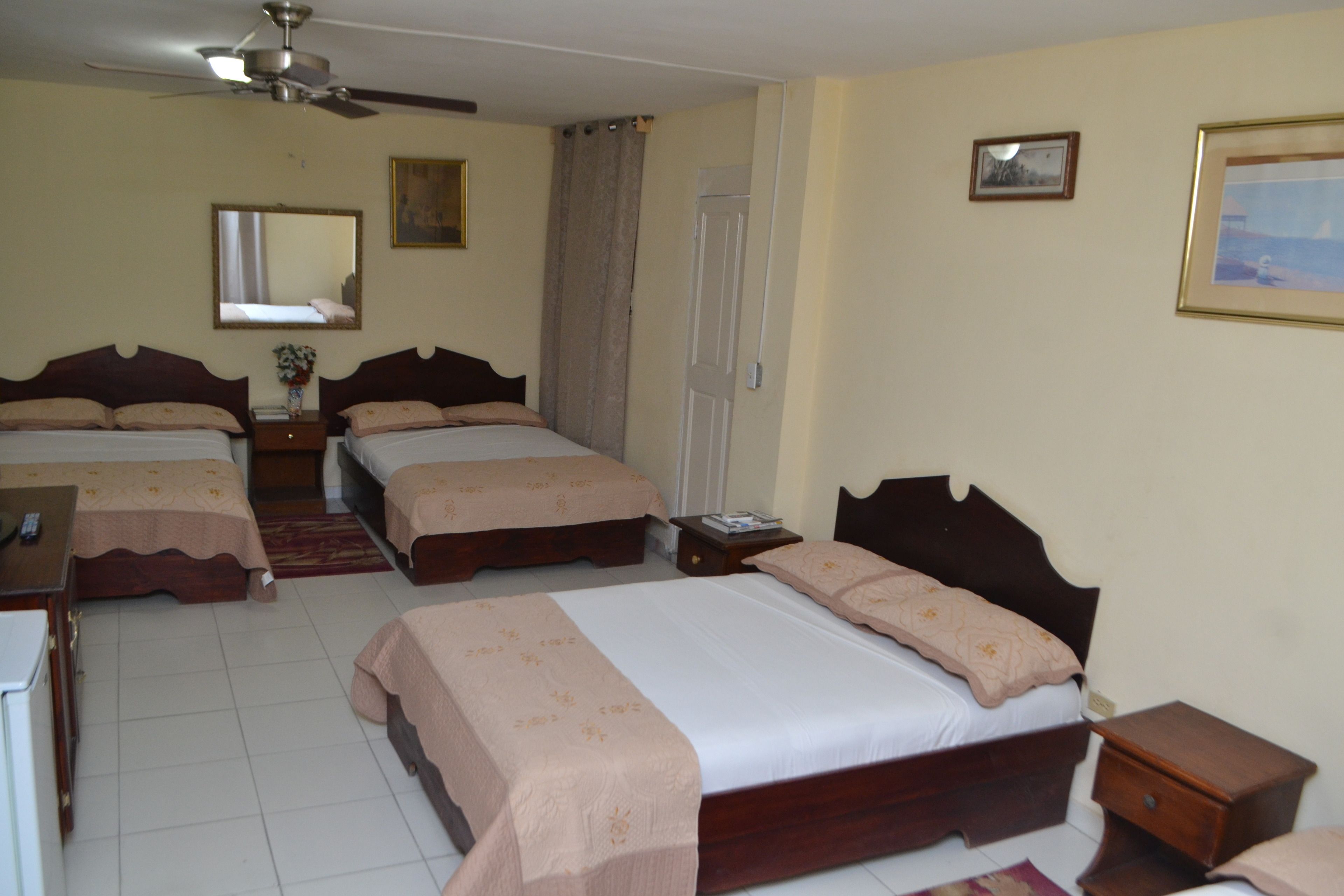 A hotel room with four single beds, a ceiling fan, a mirror, a door, and a small table with a lamp.
