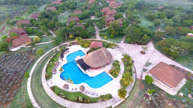 Eine Vogelperspektive zeigt ein Resort mit mehreren strohgedeckten Hütten, einem zentralen Swimmingpool und umliegenden, bepflanzten Flächen.