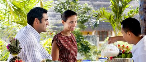 Un área de comedor al aire libre con ambiente tropical, con una mesa preparada para tres personas, rodeada de diversas frutas y un adorno central.