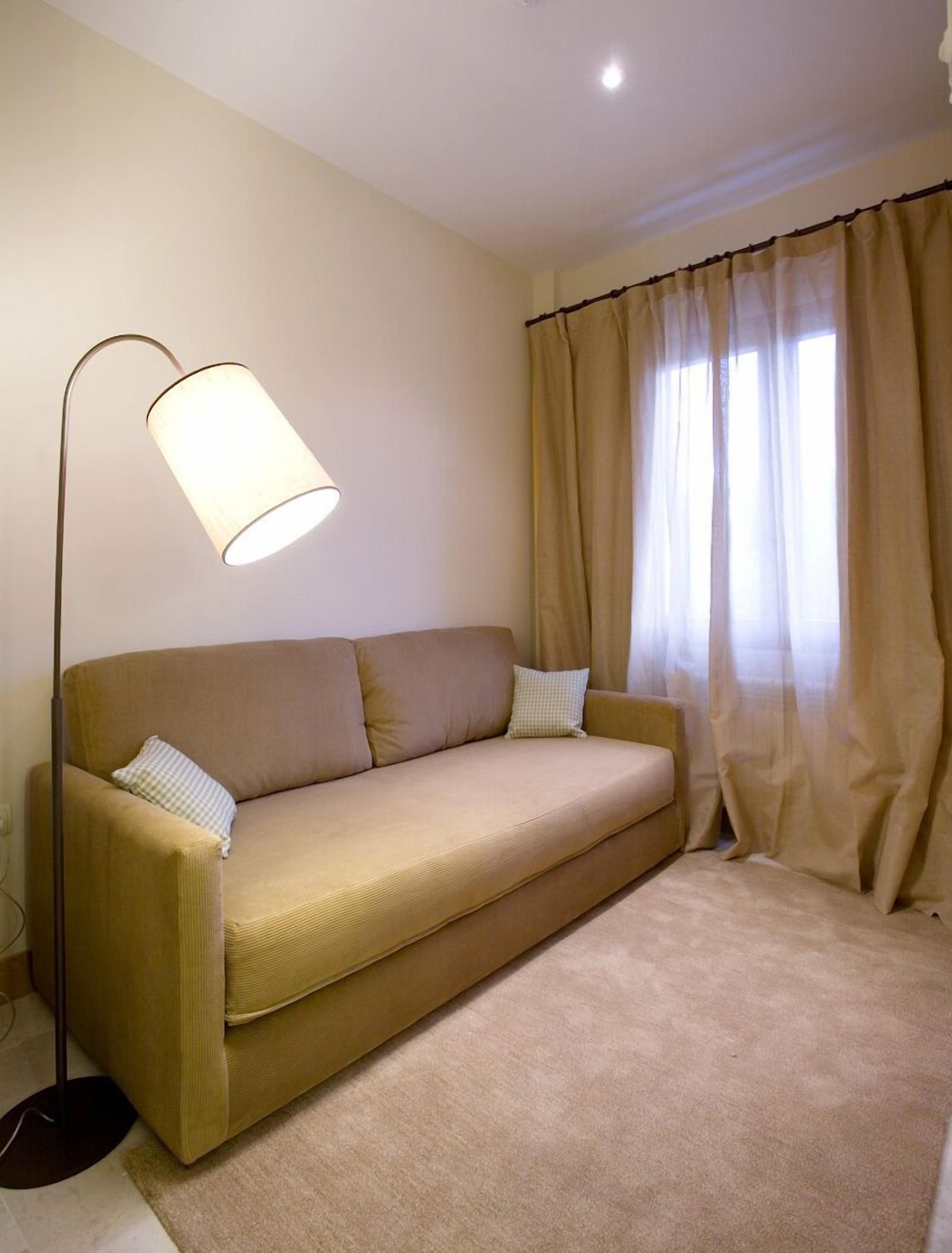 Un sofá color beige con dos almohadas, una lámpara de pie y cortinas color beige en una habitación con piso alfombrado.