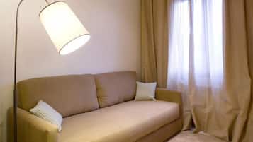Un sofá color beige con dos almohadas, una lámpara de pie y cortinas color beige en una habitación con piso alfombrado.
