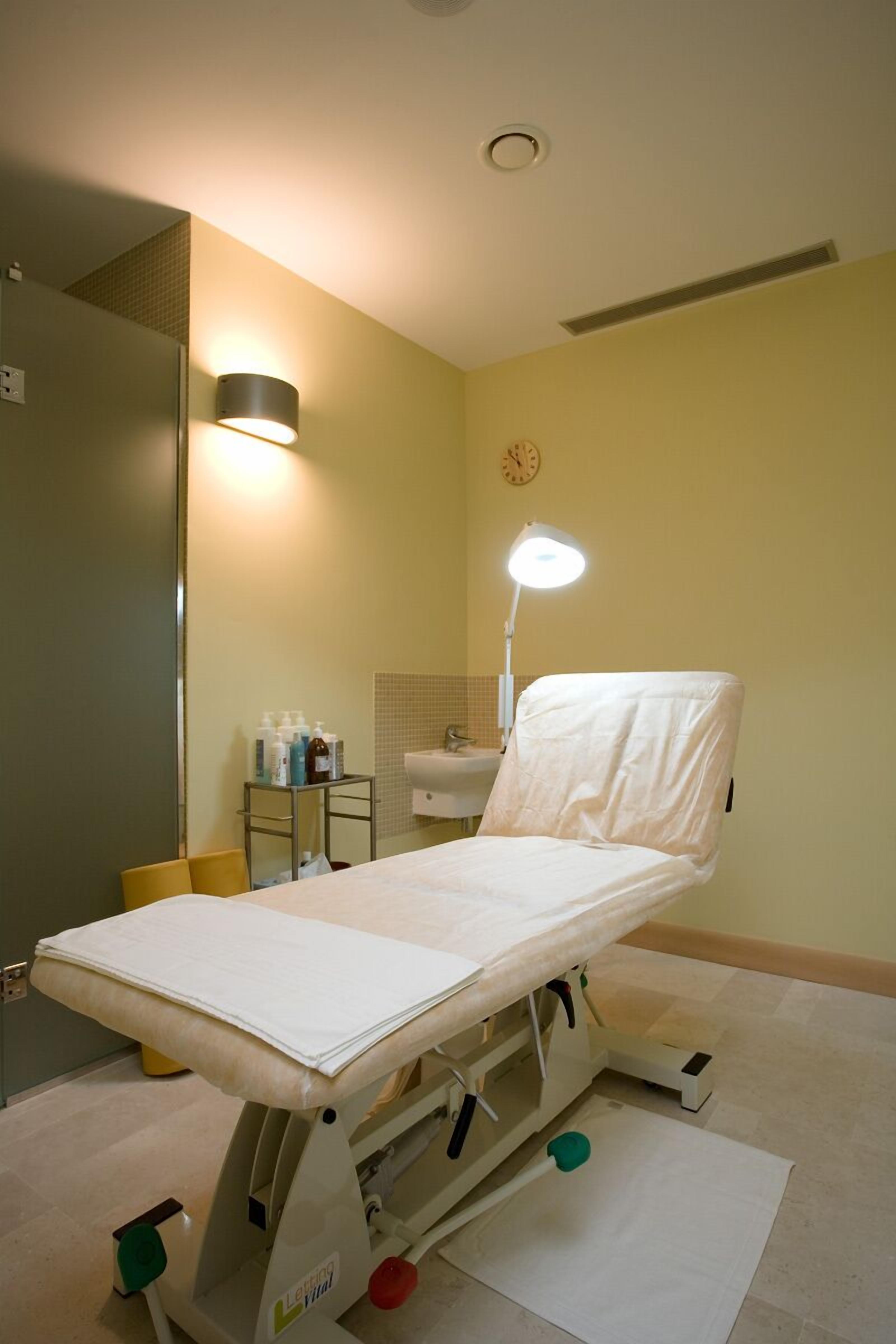 Een medisch onderzoekbed in een kamer met een spiegel, een wastafel en een klok aan de muur.