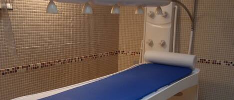 A medical examination table with a blue mat, a footrest, and a wall-mounted lamp.