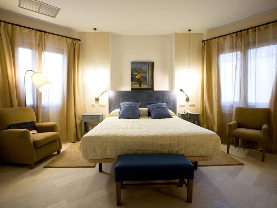 A hotel room with a large bed, two armchairs, a bench, a nightstand, a lamp, and a painting on the wall.