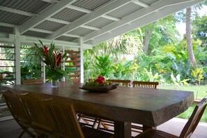 A wooden table with chairs, a bowl of fruit, and a vase of flowers in a covered outdoor area with a view of lush greenery. - Vale Vale Beachfront Villas (Port Vila)