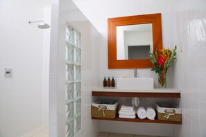 A bathroom with white tiles, a wooden-framed mirror, a white sink, and a shelf with towels and baskets. - Vale Vale Beachfront Villas (Port Vila)
